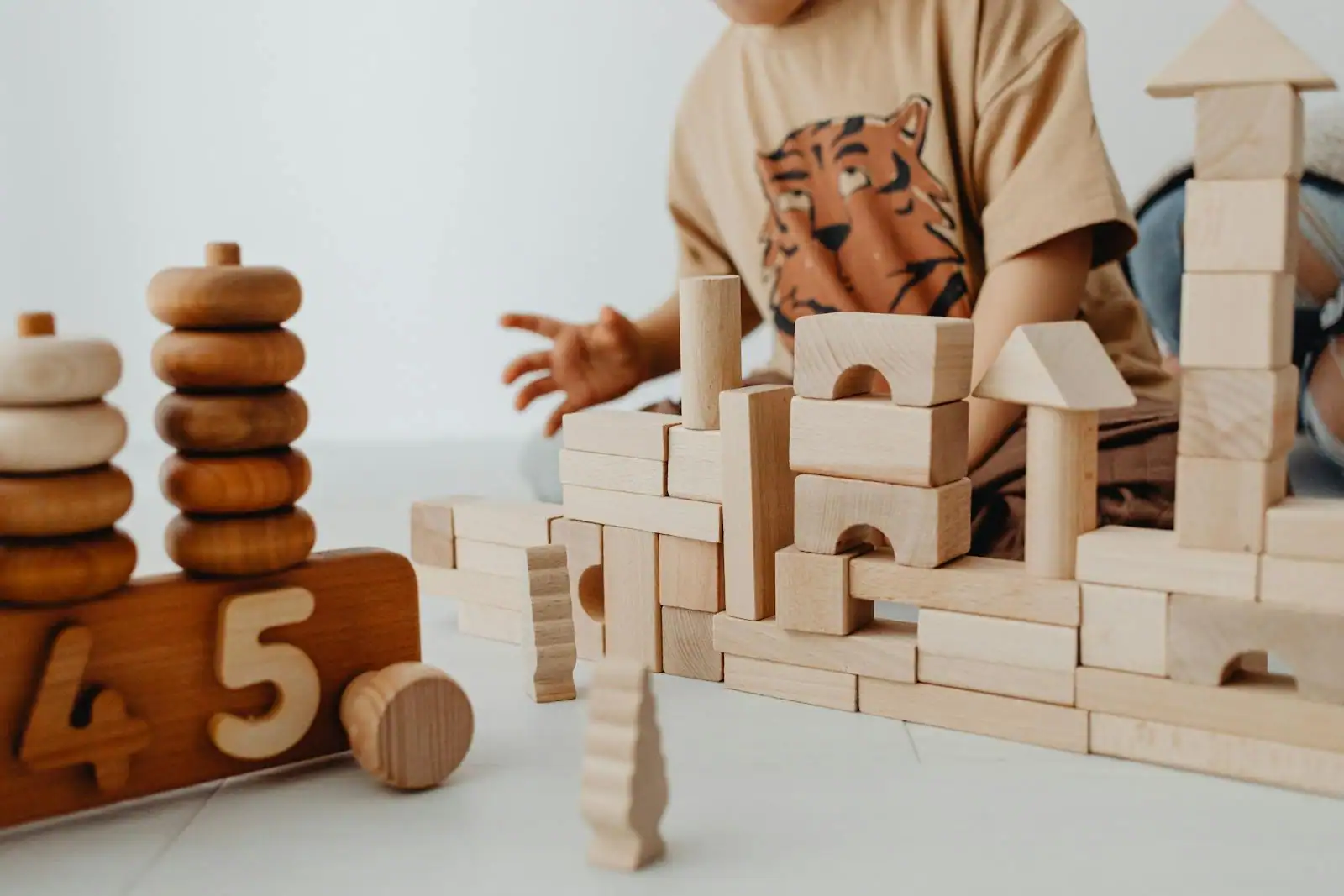Enfant construisant une tour avec des planchettes Kapla en bois clair posées en croix sur un parquet lumineux
