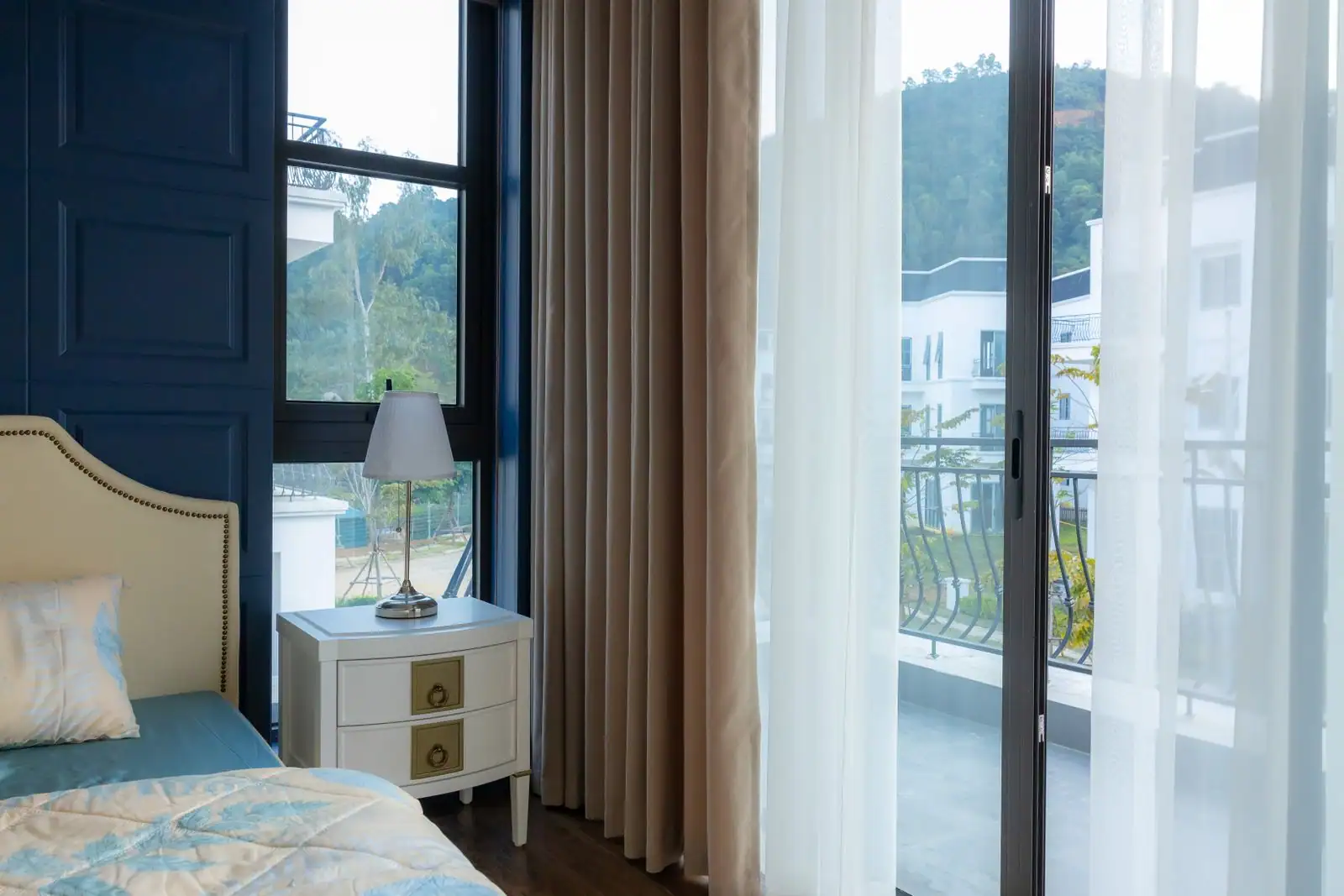 Chambre avec une grande baie vitrée et un voilage, ouvrant sur un balcon et un paysage de collines.