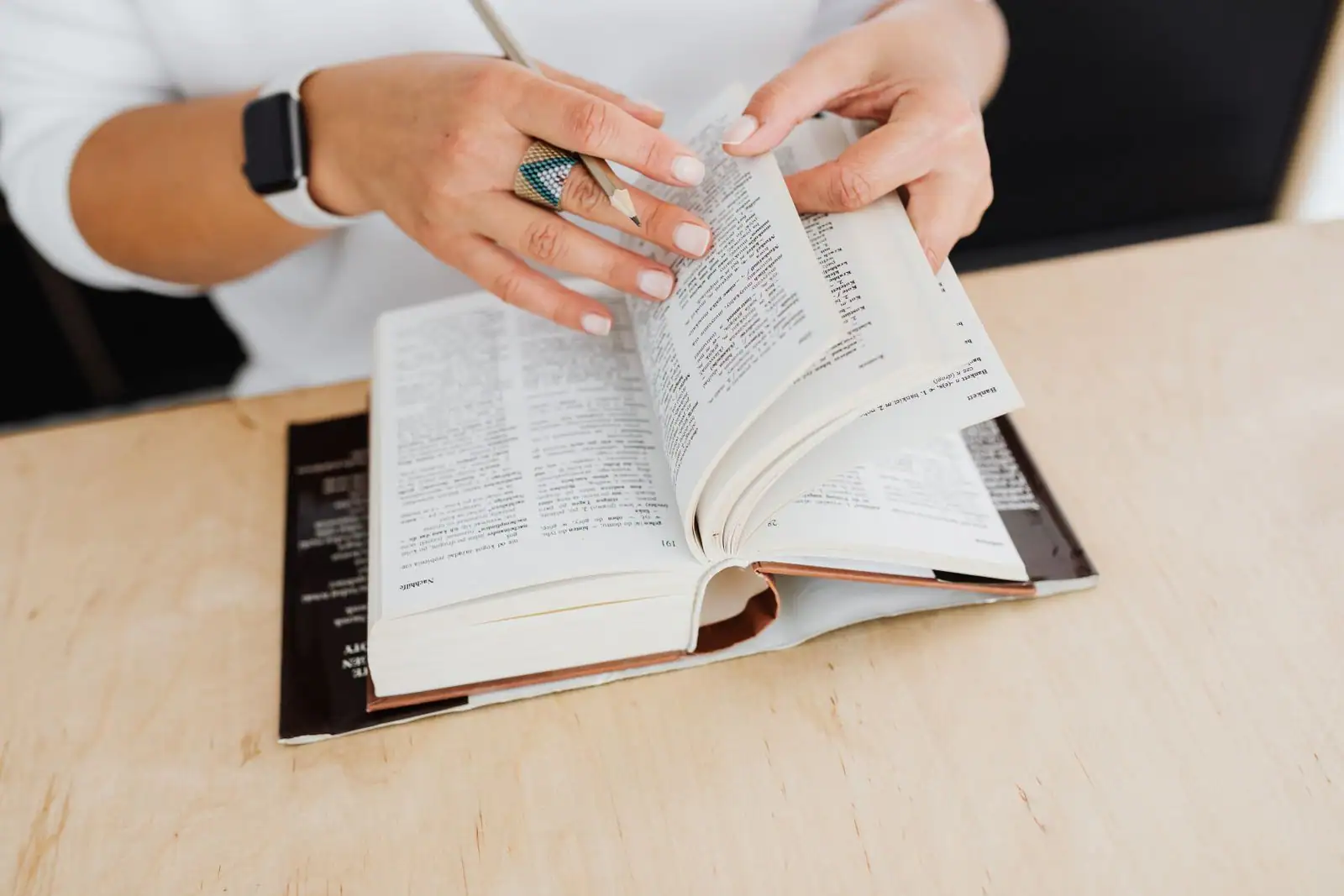 Mains feuilletant les pages d'un dictionnaire ouvert sur une table en bois, un crayon à la main