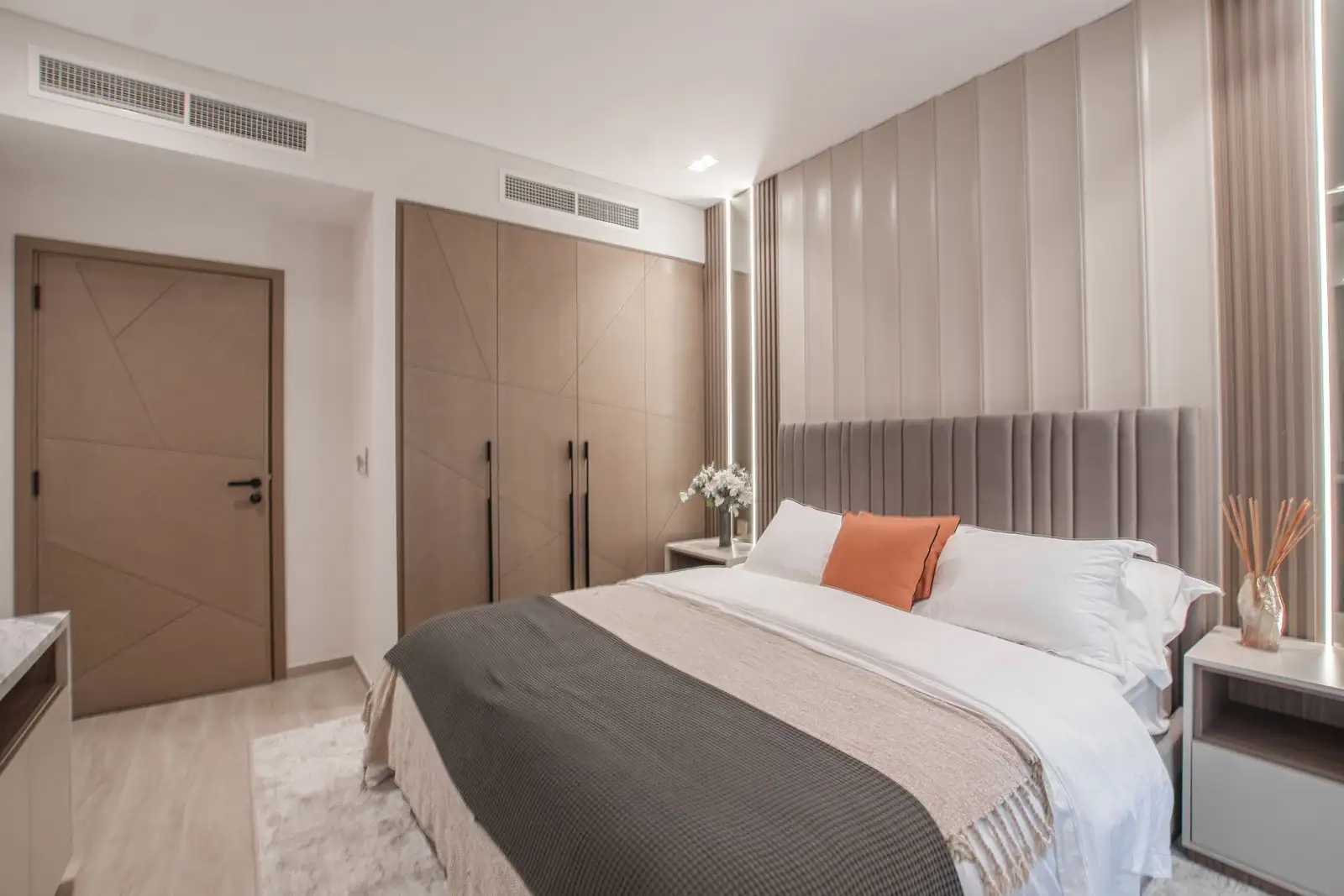 Chambre à coucher moderne aux tons neutres, avec tête de lit capitonnée grise, coussin orange et linge superposé.
