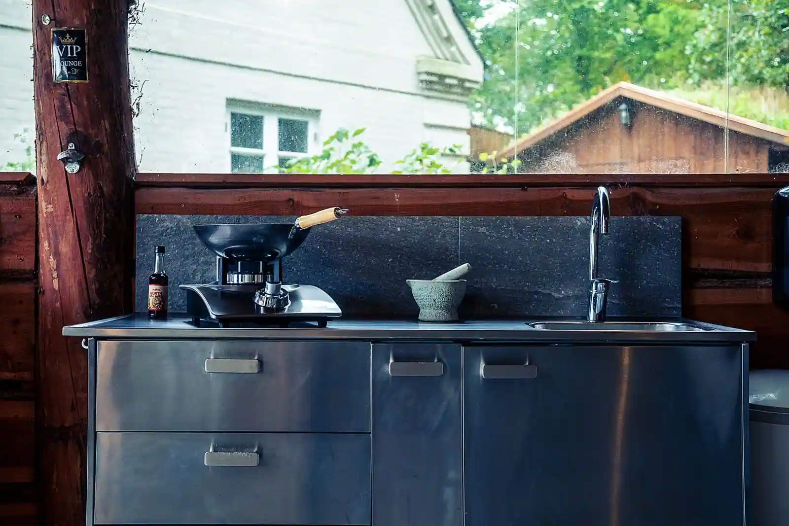 Cuisine extérieure avec meubles en inox, plan de travail en pierre et évier, sous un abri en bois
