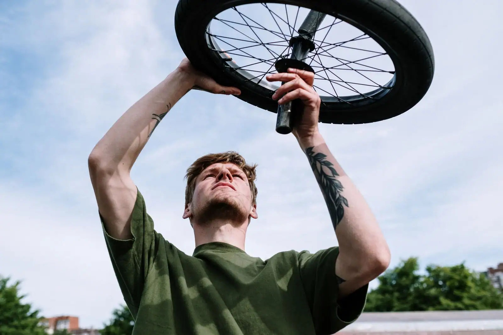 Un homme tient à bout de bras une roue de vélo et en inspecte le pneu, dehors sous un ciel clair.