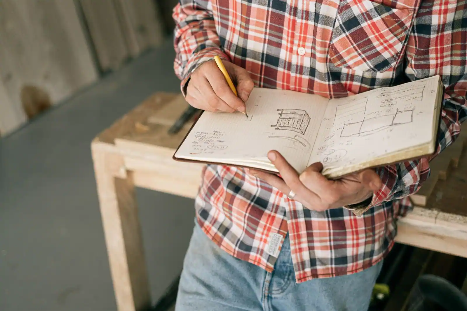 Artisan menuisier dessinant au crayon un projet de meuble dans un carnet, au milieu de son atelier