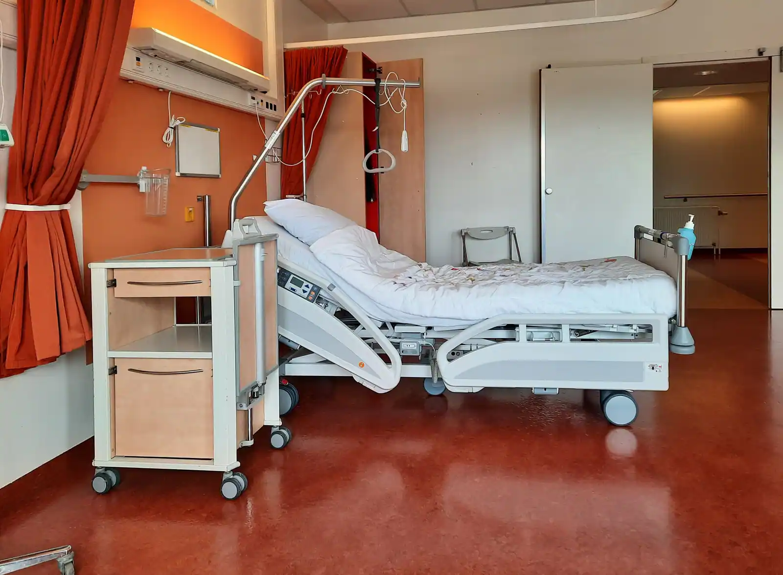 Chambre d'hôpital inoccupée, lit médicalisé relevé et table de chevet ouvrant sur le couloir