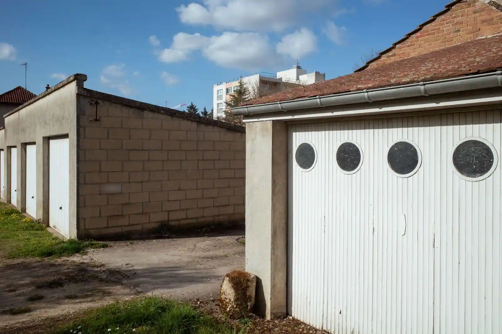 Rangée de garages individuels en parpaing, portes basculantes blanches fermées, sur une cour goudronnée.